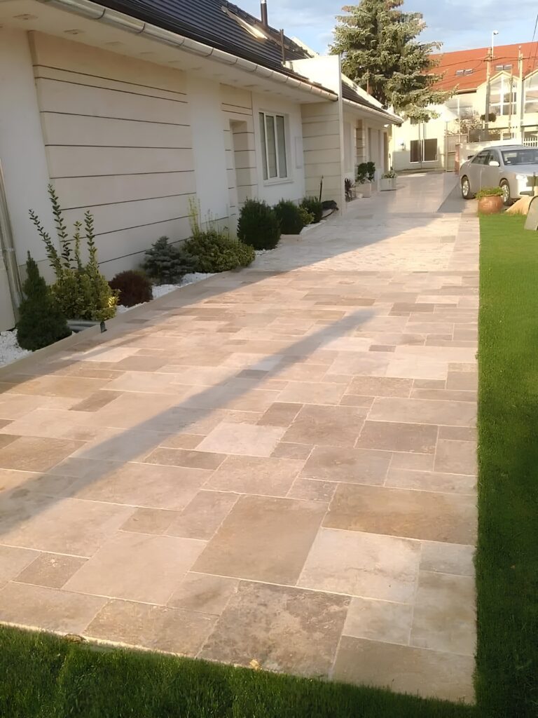 Travertin pour allée : Dallage extérieur et jardin moderne. Allée en dalles de travertin clair, bordée d'une pelouse verte et d'arbustes taillés. Longue façade blanche d'une maison moderne.