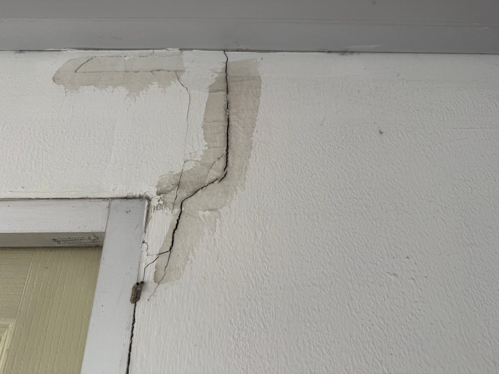 Fissure profonde sur mur : signes de dégât des eaux au plafond. Fissure majeure sur mur blanc, partant du plafond au-dessus d'un cadre de porte. Taches grises révélant un dégât des eaux et une tentative de réparation.