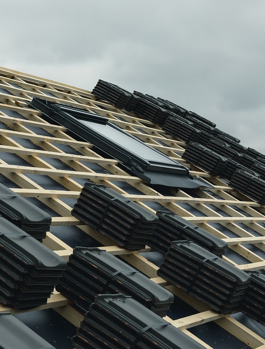 Pose de toiture neuve : Tuiles noires et fenêtre de toit. Toit en construction : charpente, sous-couche noire, fenêtre de toit et piles de tuiles foncées prêtes à la pose.